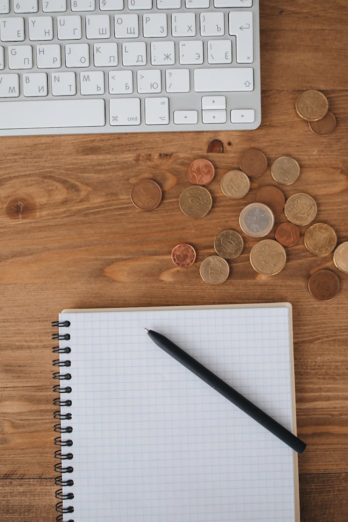Overhead view of a workspace with keyboard, coins, and a notebook on a wooden desk